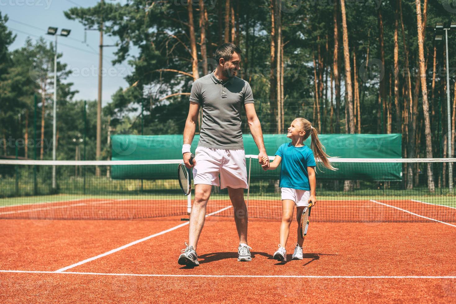 toute la longueur de la petite fille aux cheveux blonds en vêtements de sport portant une raquette de tennis et regardant son père marchant près d'elle par un court de tennis photo