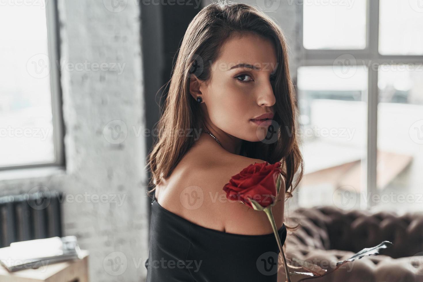 beauté sans effort. jolie jeune femme en robe noire élégante tenant une rose rouge tout en passant du temps à la maison photo