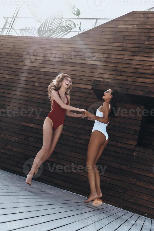 danse spontanée. toute la longueur de deux jolies jeunes femmes en maillot de bain souriant tout en dansant contre le mur en bois à l'extérieur photo