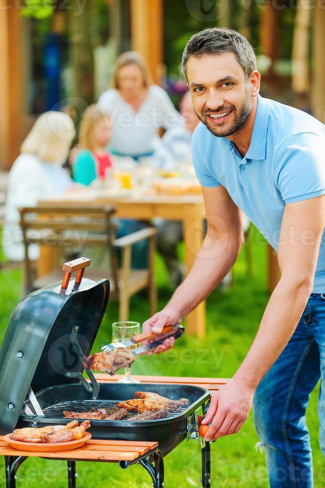 profiter d'un barbecue en famille. heureux jeune homme faisant griller de la viande sur le gril et souriant tandis que d'autres membres de la famille assis à la table à manger en arrière-plan photo