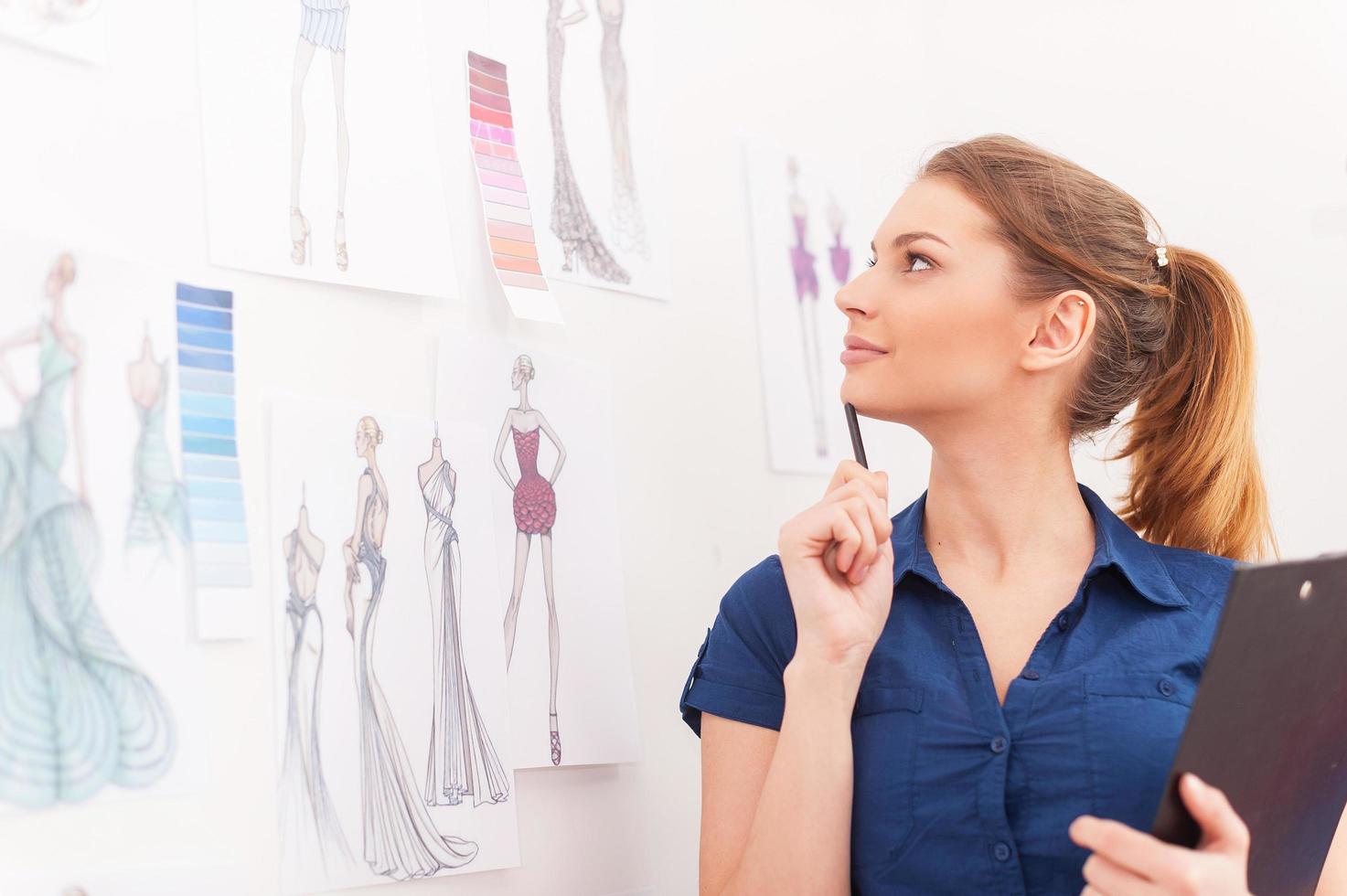 créer un nouveau look de mode. belle créatrice de mode féminine tenant le presse-papiers et regardant les croquis accrochés au mur photo