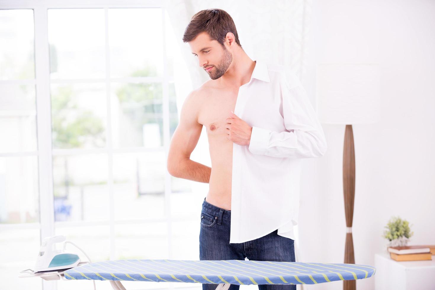 portant sa chemise préférée. jeune homme confiant habillant une chemise blanche après le repassage tout en se tenant près de la planche à repasser photo