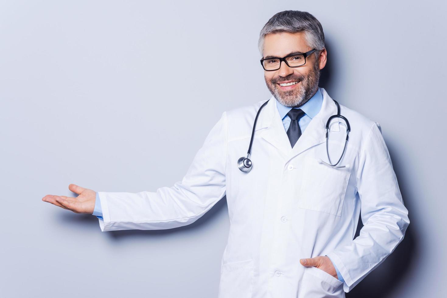 médecin faisant la publicité de votre produit. joyeux médecin mature regardant la caméra et pointant vers l'extérieur en se tenant debout sur fond gris photo