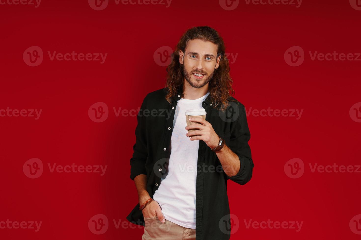 charmant jeune homme en vêtements décontractés regardant la caméra et souriant debout sur fond rouge photo