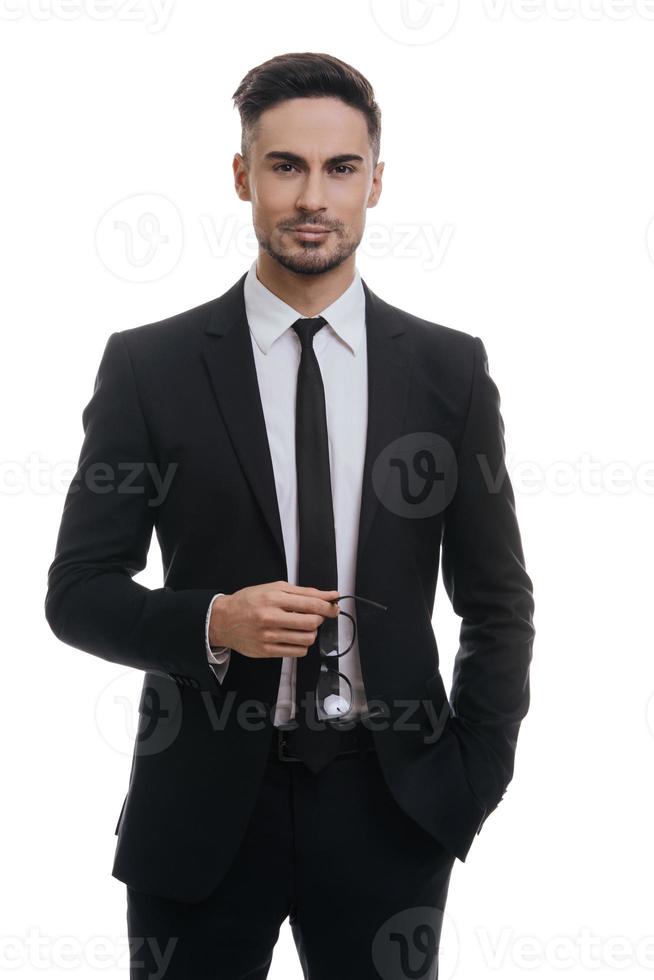 meilleur gestionnaire de tous les temps. beau jeune homme en costume complet tenant des lunettes et regardant la caméra avec le sourire en se tenant debout sur fond blanc photo