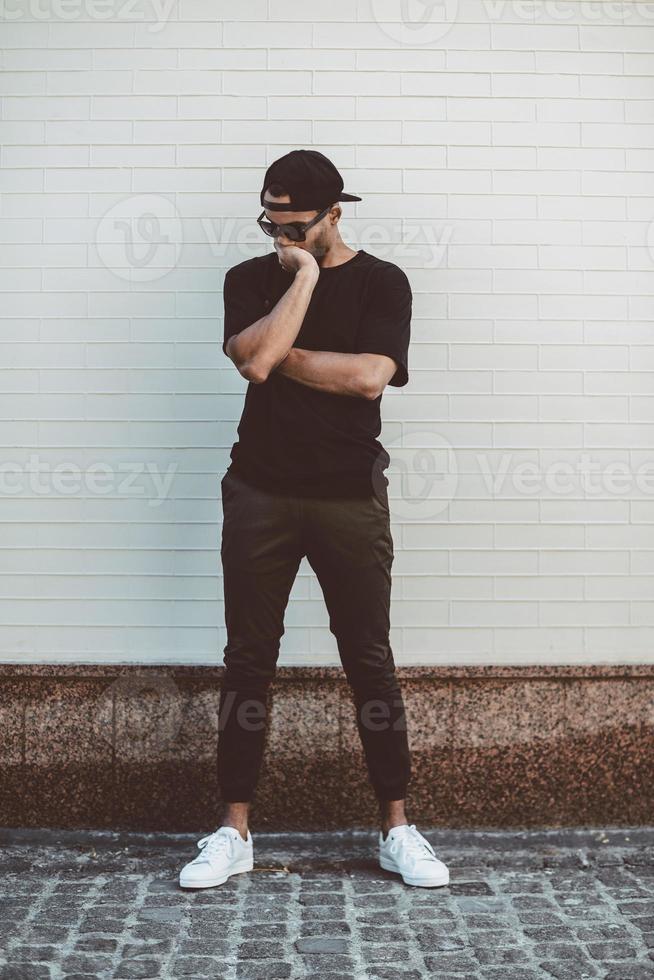 perdu dans les pensées. toute la longueur du jeune homme de race mixte en vêtements décontractés debout devant le mur de briques et couvrant le visage avec la main photo