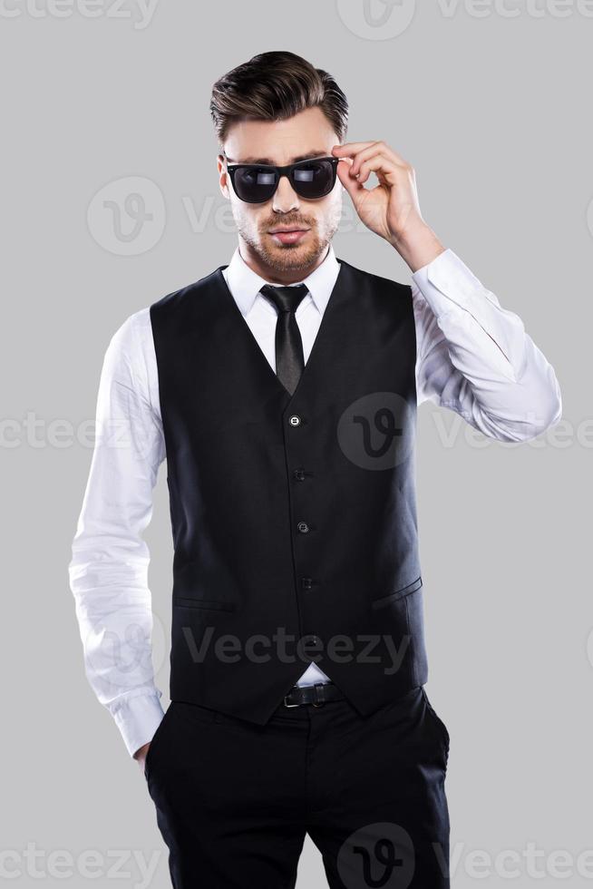élégant et beau. beau jeune homme en tenue de soirée ajustant ses lunettes de soleil et regardant la caméra en se tenant debout sur fond gris photo