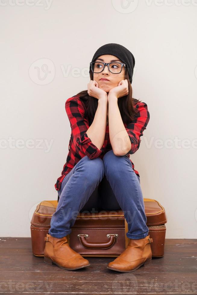 s'ennuyer. belle jeune femme en couvre-chef tenant les mains sur le menton et regardant loin tout en étant assis sur la valise photo