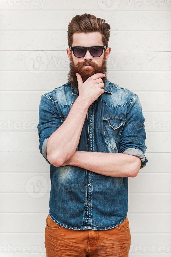 cool et confiant. élégant jeune homme barbu regardant la caméra et tenant la main sur le menton tout en se tenant à l'extérieur photo