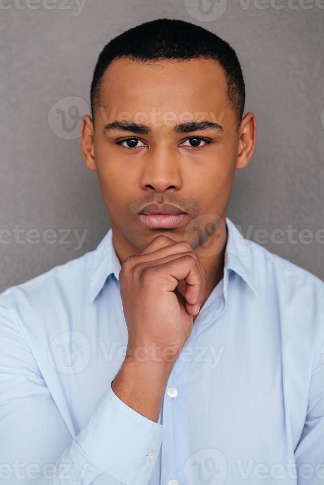 portrait de confiance. confiant jeune homme africain tenant la main sur le menton et regardant la caméra en se tenant debout sur fond gris photo