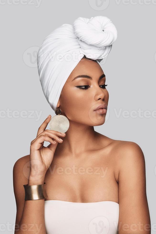 dangereusement belle. séduisante jeune femme africaine en turban ajustant la boucle d'oreille et regardant loin en se tenant debout sur fond gris photo