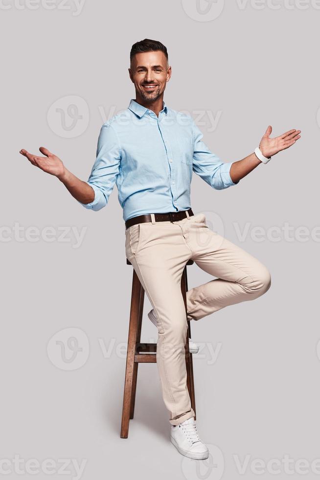 salutation. toute la longueur d'un beau jeune homme souriant et gardant les bras tendus assis sur un tabouret sur fond gris photo