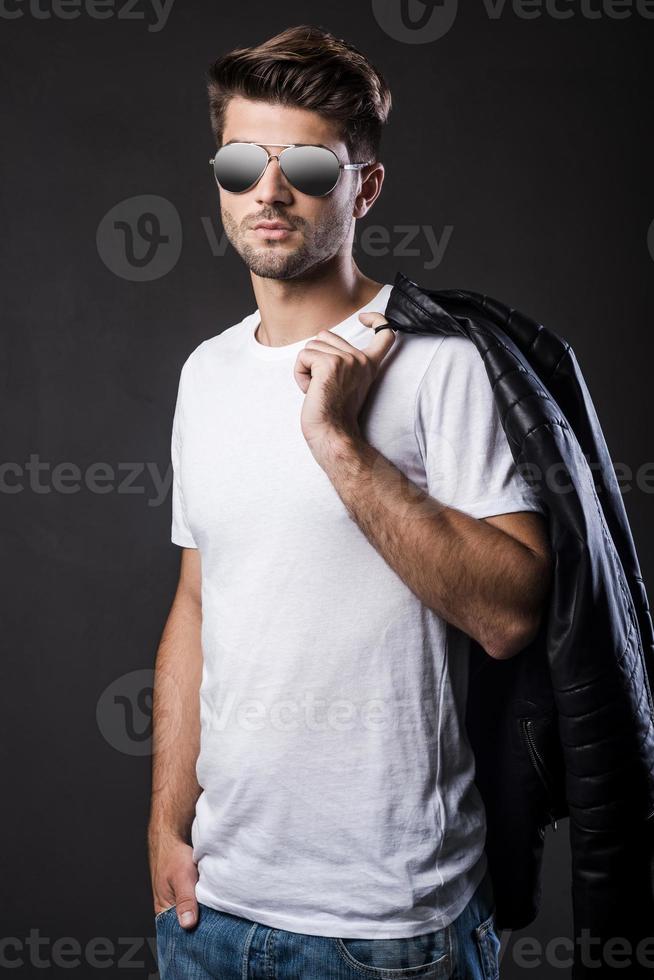 beau à la mode. beau jeune homme à lunettes de soleil portant sa veste en cuir sur l'épaule et regardant la caméra en se tenant debout sur fond noir photo