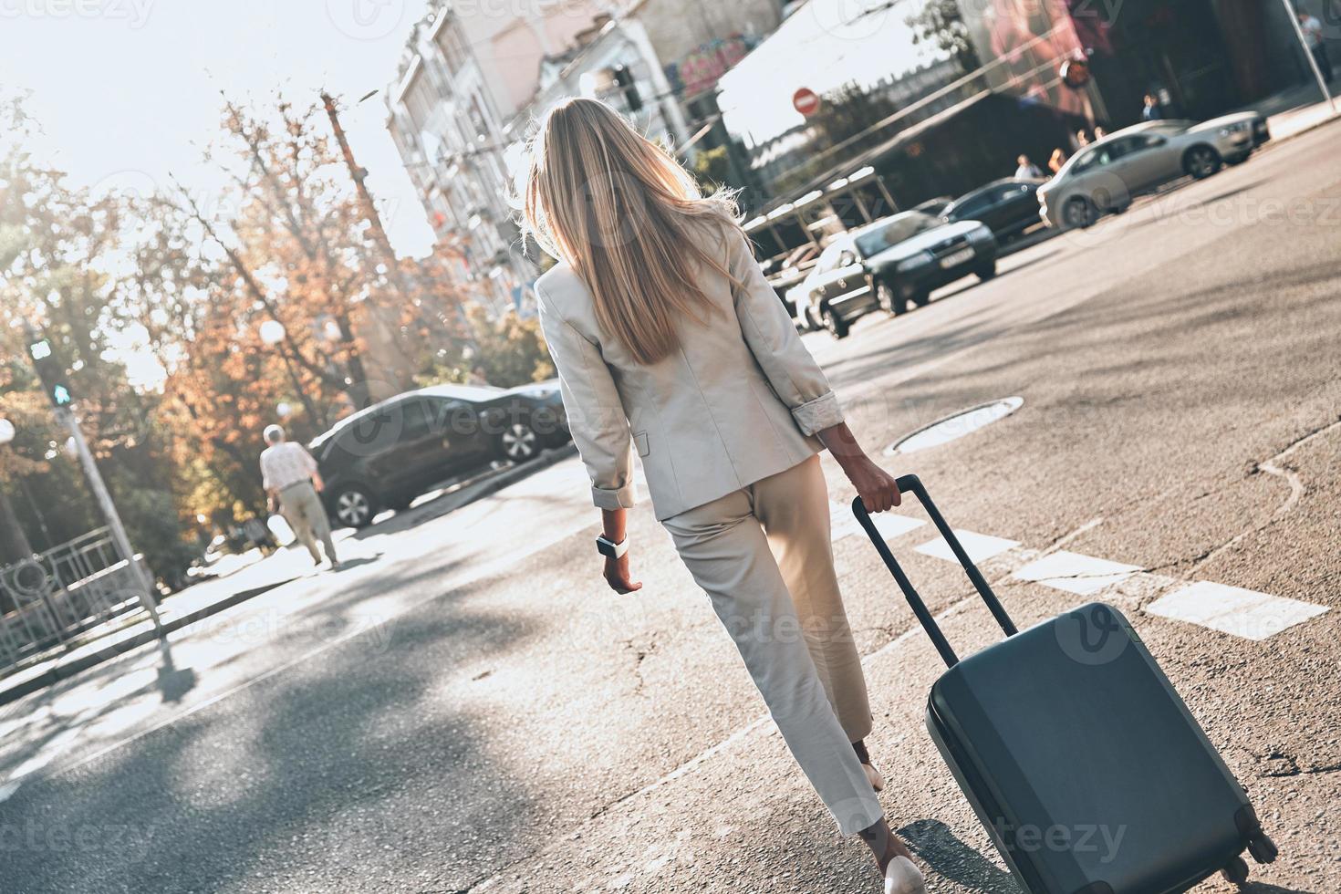 en route vers de nouvelles opportunités. vue arrière de la jeune femme en costume tirant des bagages tout en marchant à l'extérieur photo