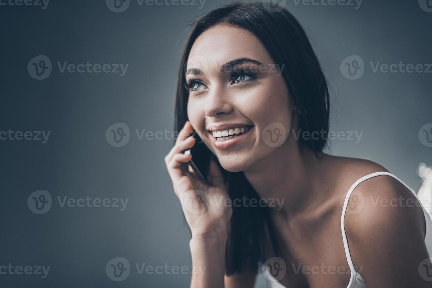 bonne conversation avec un ami. jolie jeune femme parlant au téléphone mobile et souriant assis contre un mur gris photo