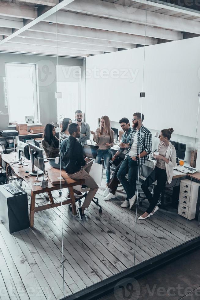 groupe de jeunes professionnels. vue de dessus pleine longueur de jeunes gens modernes en vêtements décontractés intelligents discutant de quelque chose tout en se tenant derrière le mur de verre dans le bureau de création photo