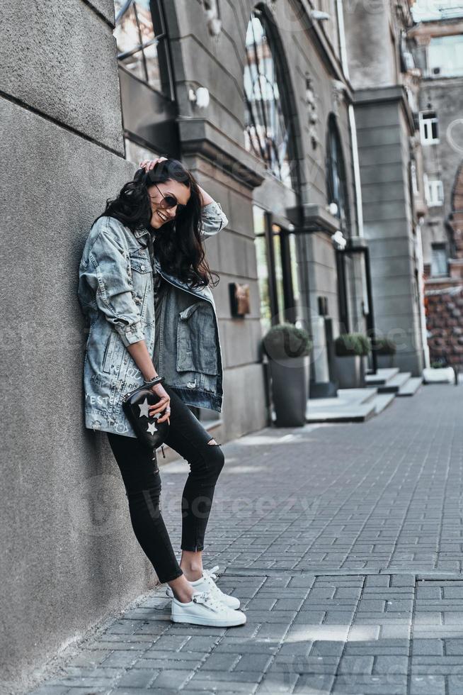 style de rue. toute la longueur de la belle jeune femme en veste en jean gardant la main dans les cheveux tout en s'appuyant sur le bâtiment à l'extérieur photo