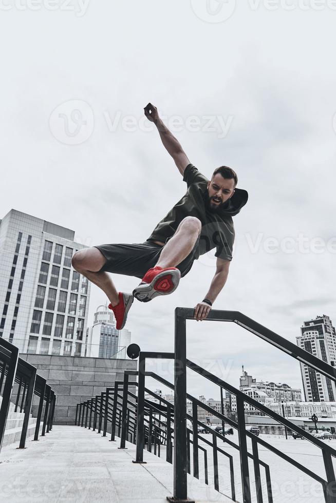 sauter directement par-dessus tous les obstacles. toute la longueur du beau jeune homme en vêtements de sport sautant par-dessus la balustrade tout en faisant de l'exercice à l'extérieur photo