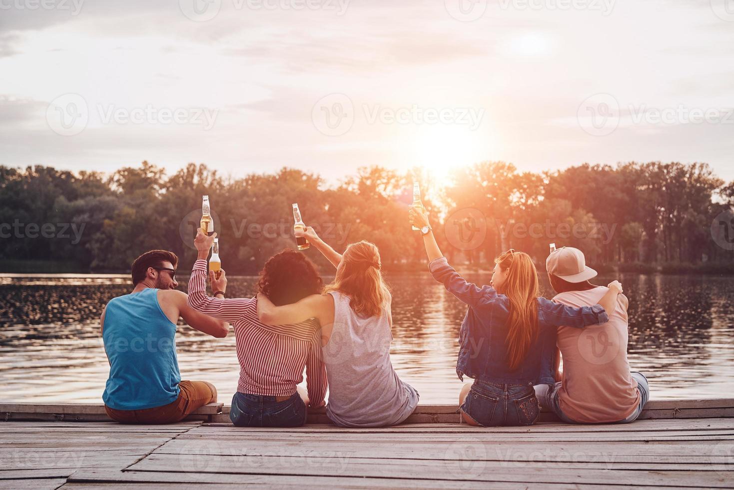 vue arrière de jeunes en tenue décontractée portant un toast avec des bouteilles de bière assis sur la jetée photo