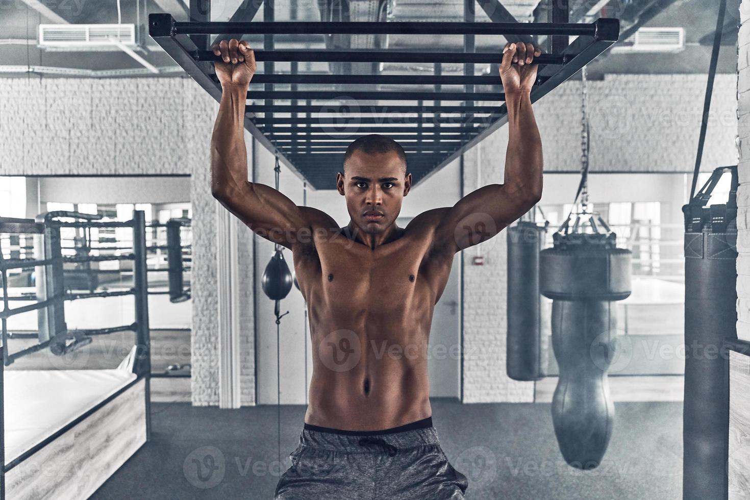 déterminé à gagner. jeune homme africain torse nu regardant la caméra et faisant des tractions tout en faisant de l'exercice dans la salle de gym photo