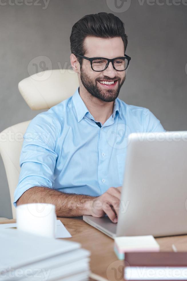 excellente solution. gros plan joyeux jeune bel homme portant des lunettes travaillant sur un ordinateur portable et souriant alors qu'il était assis sur son lieu de travail photo