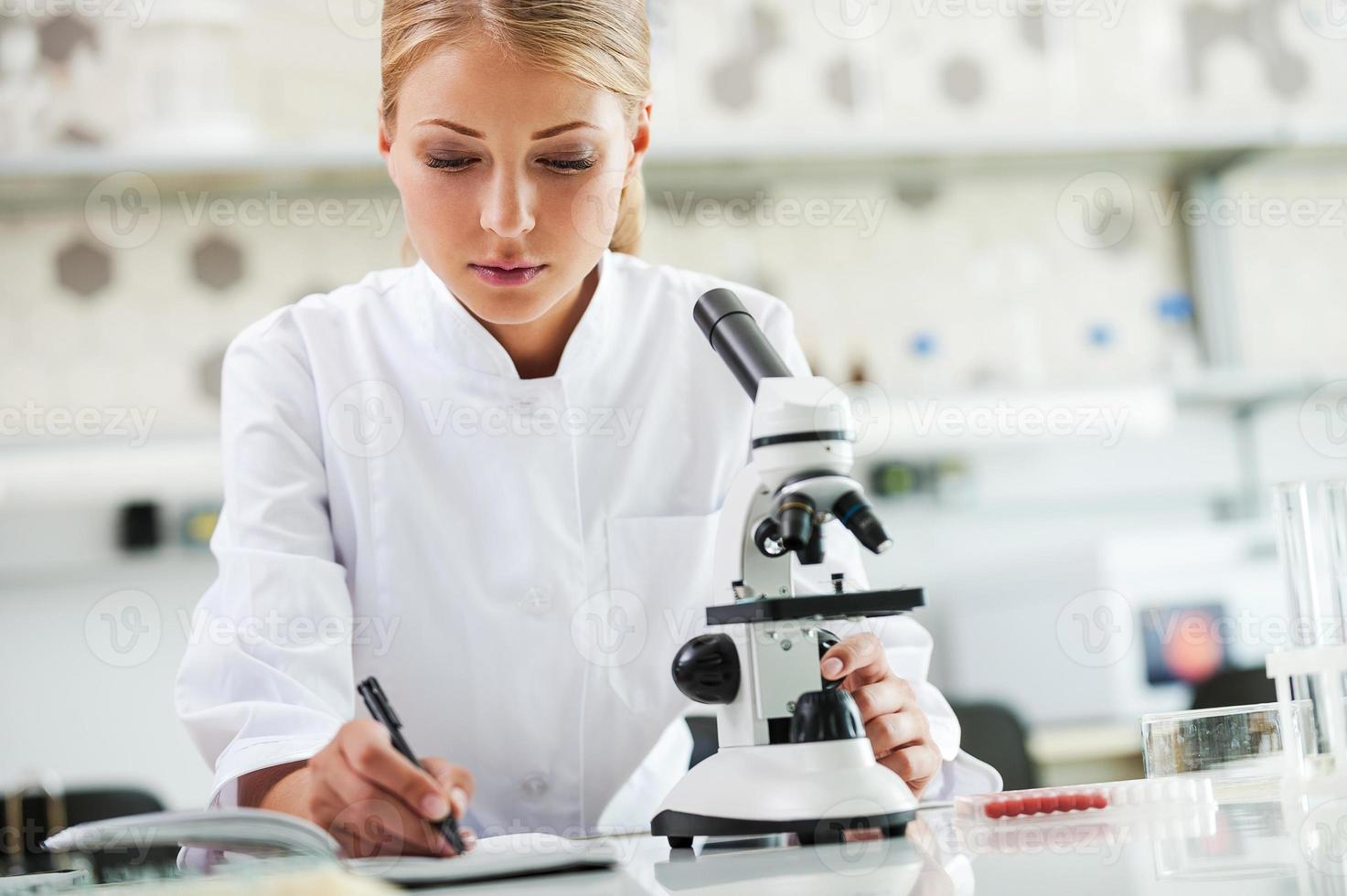 vérification des résultats. jeune femme scientifique sérieuse utilisant un microscope et écrivant dans un bloc-notes assise sur son lieu de travail photo