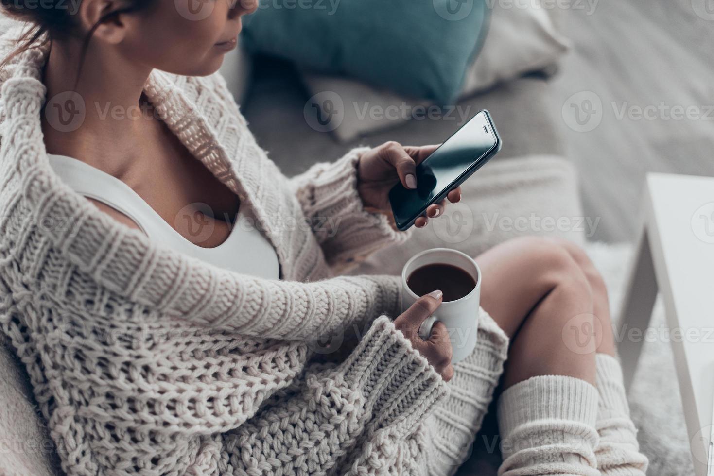 envoyer des SMS à son petit ami. gros plan d'une belle jeune femme tenant une tasse et utilisant un téléphone intelligent tout en se relaxant sur un canapé à la maison photo
