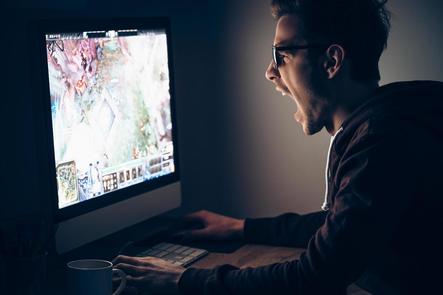 la victoire est proche. vue latérale d'un jeune homme jouant à un jeu vidéo assis à la table dans une pièce sombre photo