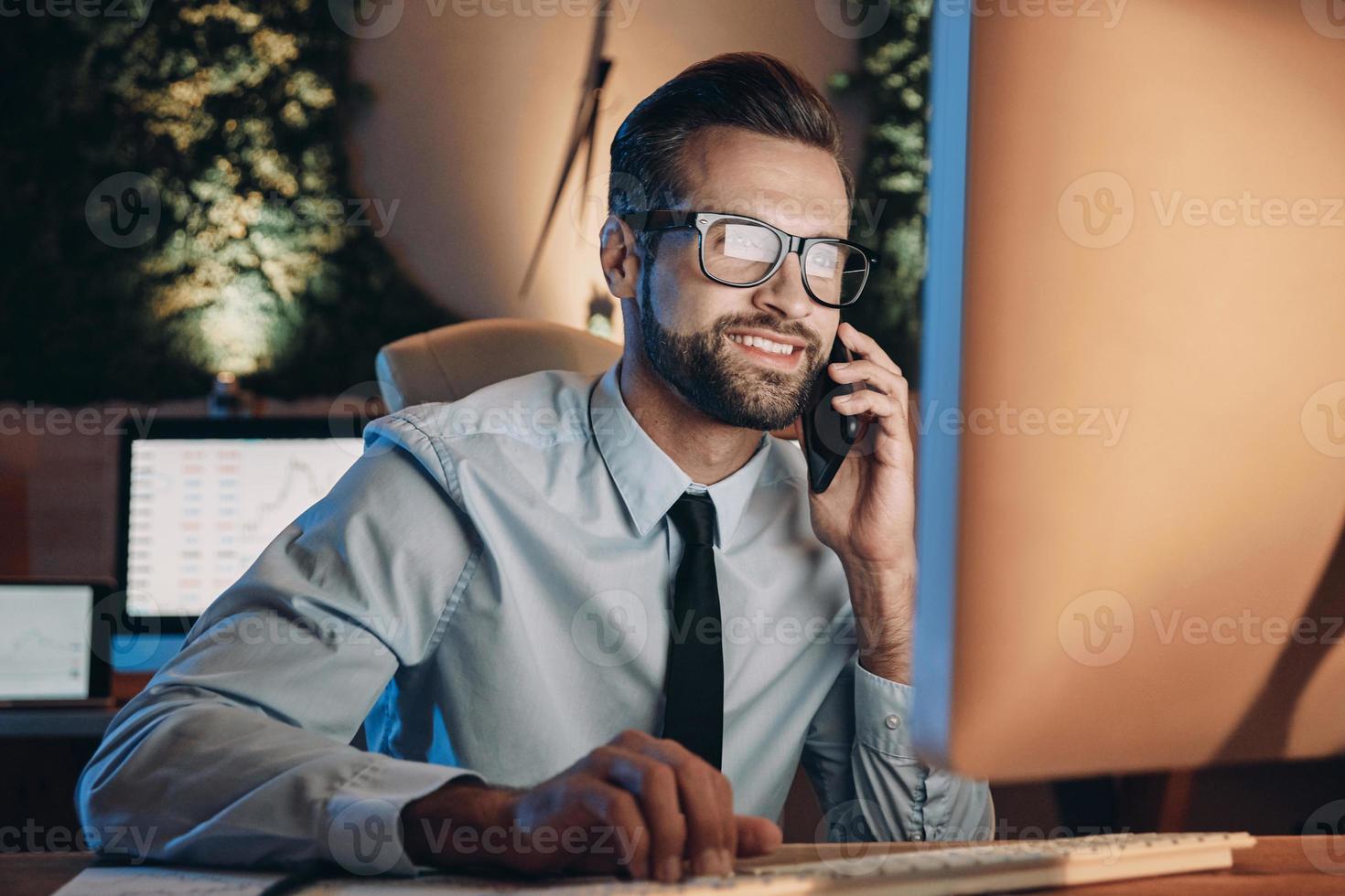 joyeux jeune homme travaillant sur ordinateur et parlant sur téléphone portable tout en restant tard au bureau photo