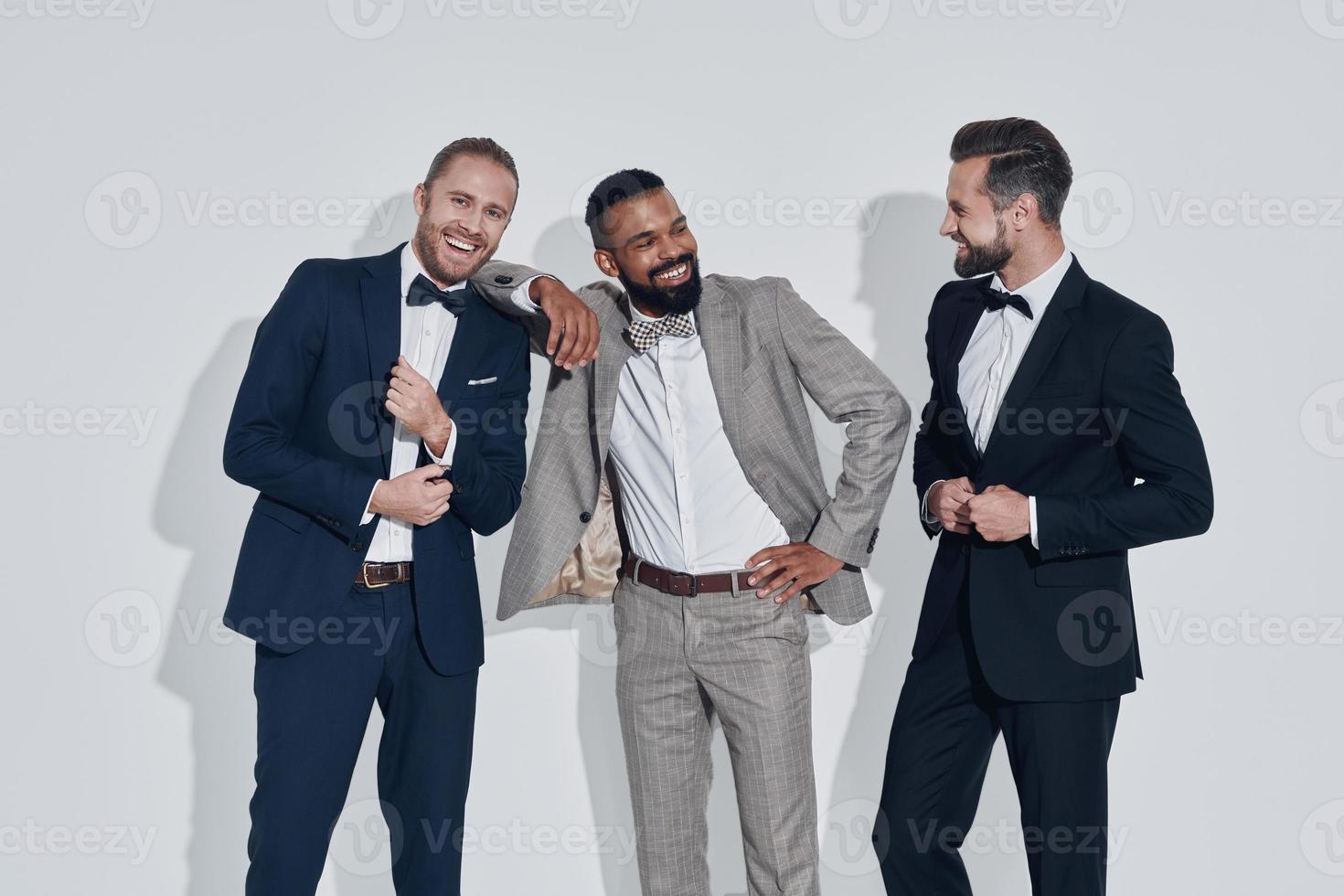 trois beaux jeunes hommes en costumes et noeuds papillon regardant la caméra et souriant en se tenant debout sur fond gris photo