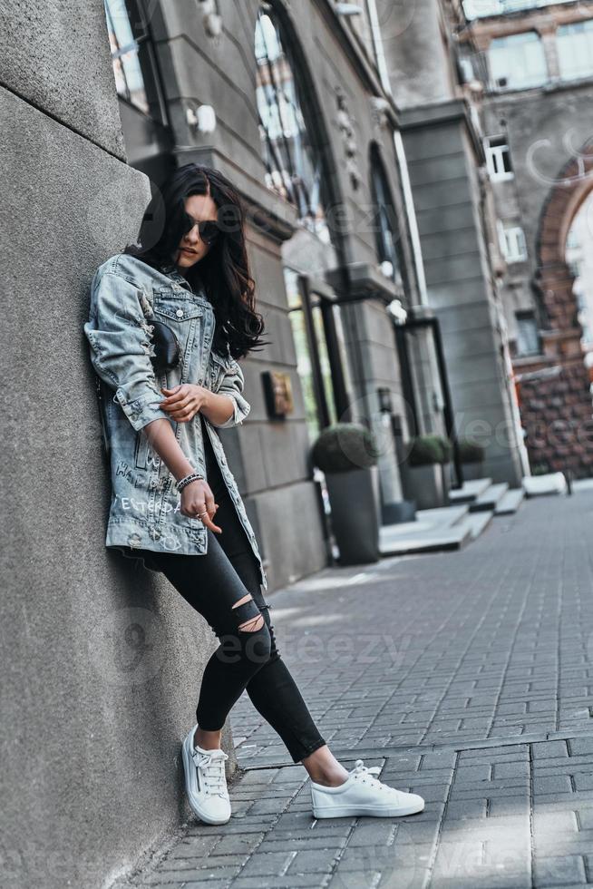 confiante dans son style. toute la longueur de la belle jeune femme en veste en jean regardant la caméra tout en s'appuyant sur le bâtiment à l'extérieur photo