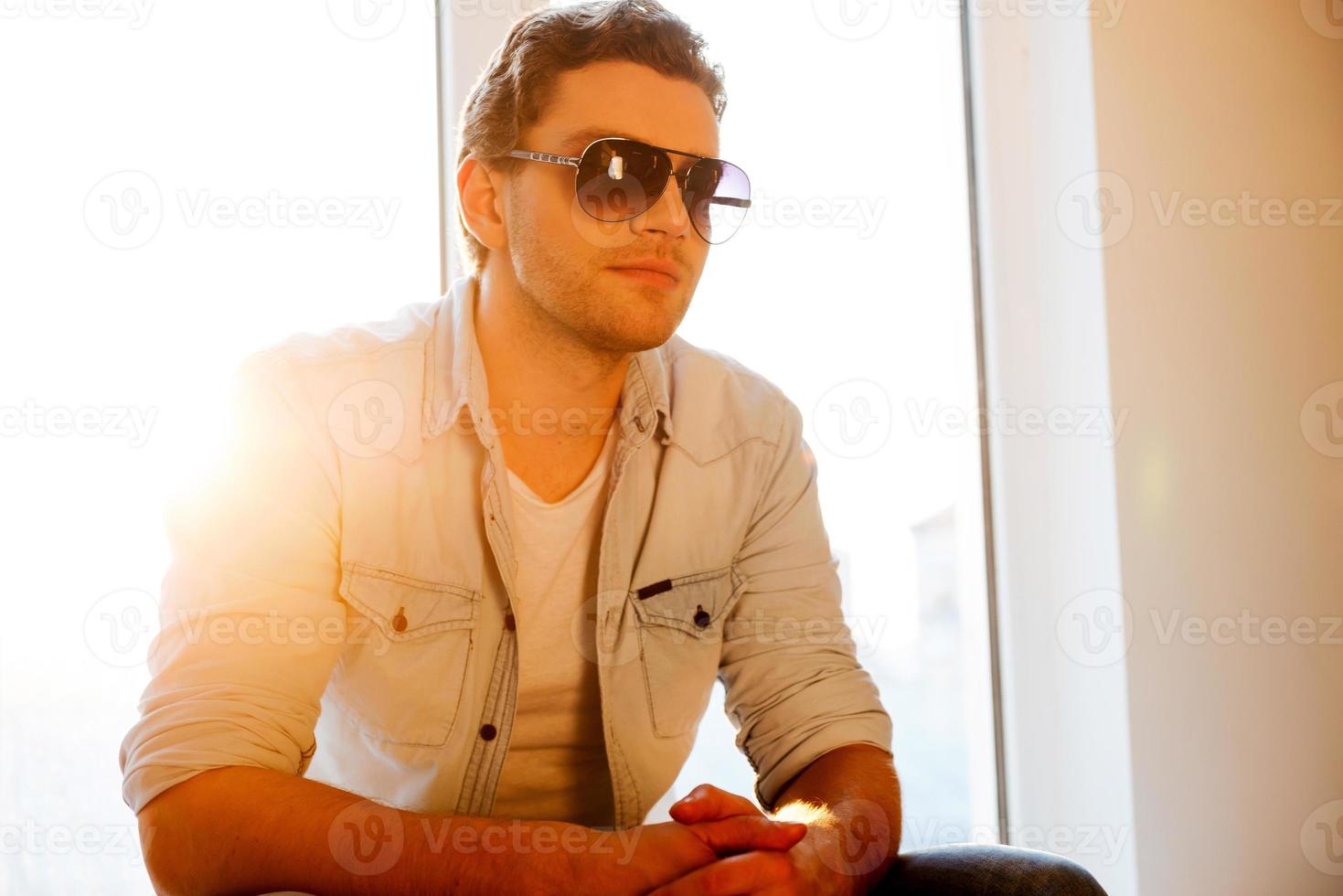 prendre le temps de se détendre. beau jeune homme à lunettes de soleil assis sur le rebord de la fenêtre en détournant les yeux photo