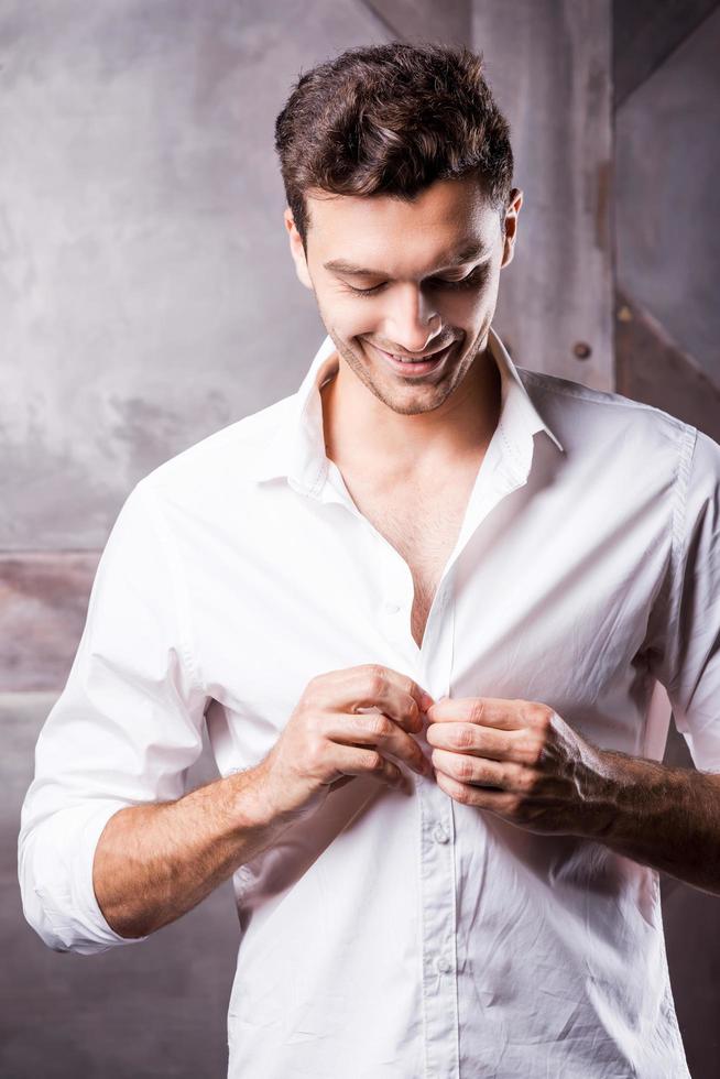 portant sa chemise préférée. beau jeune homme boutonnant sa chemise blanche et souriant photo