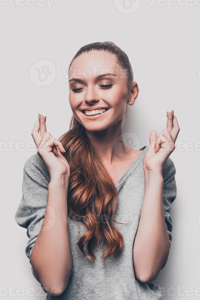 en attente d'un moment spécial. séduisante jeune femme souriante en gardant les doigts croisés et les yeux fermés en se tenant debout sur fond gris photo