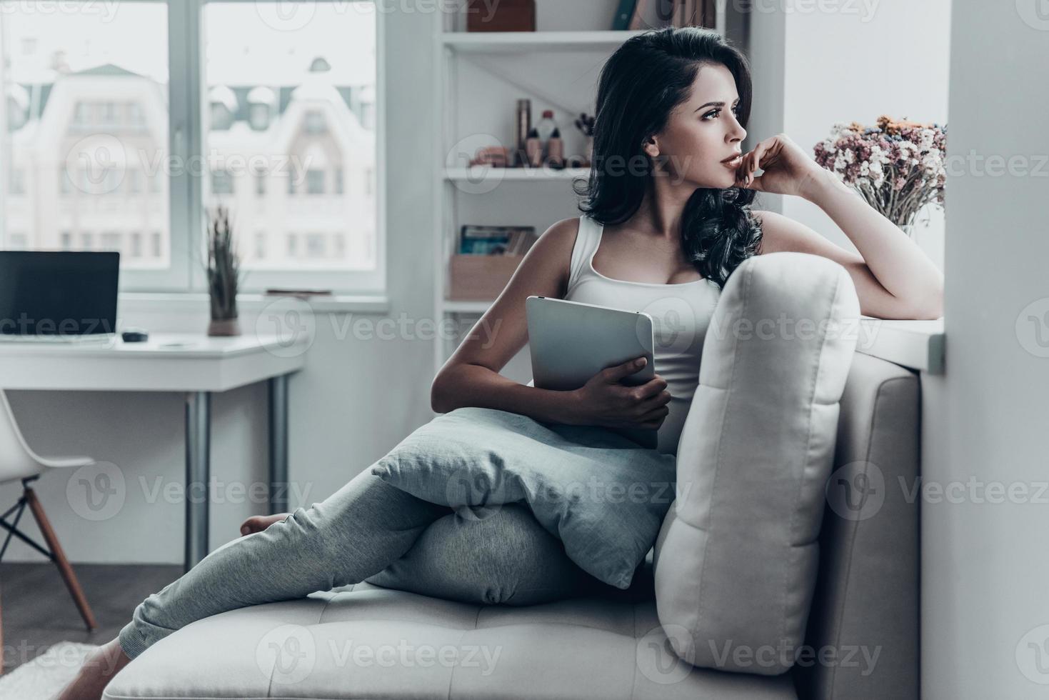 jour de flemmardise. jeune femme réfléchie tenant une tablette numérique et assise sur un canapé tout en regardant dehors par la fenêtre photo