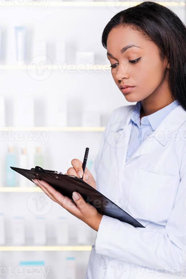 pharmacien en pharmacie. confiante jeune femme africaine en blouse de laboratoire prenant des notes dans le presse-papiers tout en se tenant dans la pharmacie photo