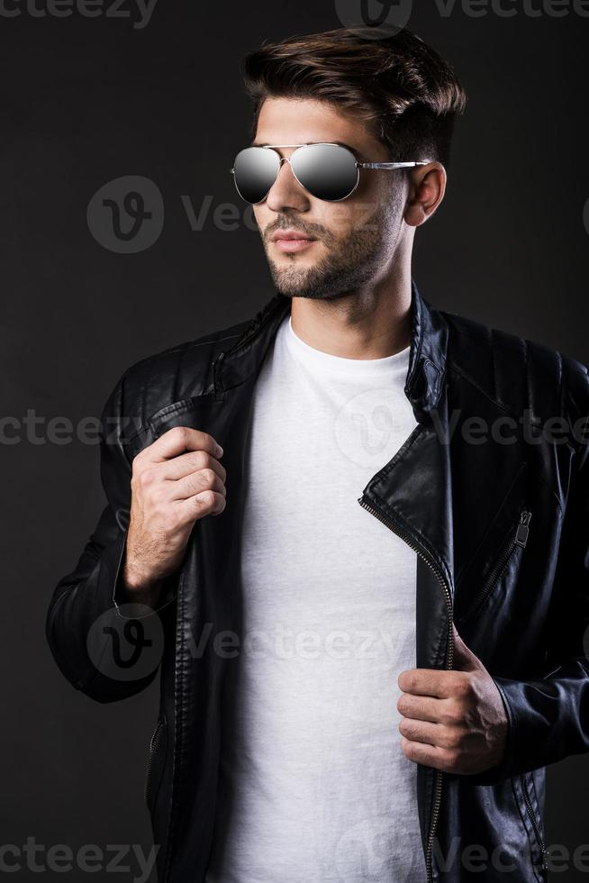 cool et tendance. beau jeune homme ajustant sa veste en cuir et regardant ailleurs en se tenant debout sur fond noir photo