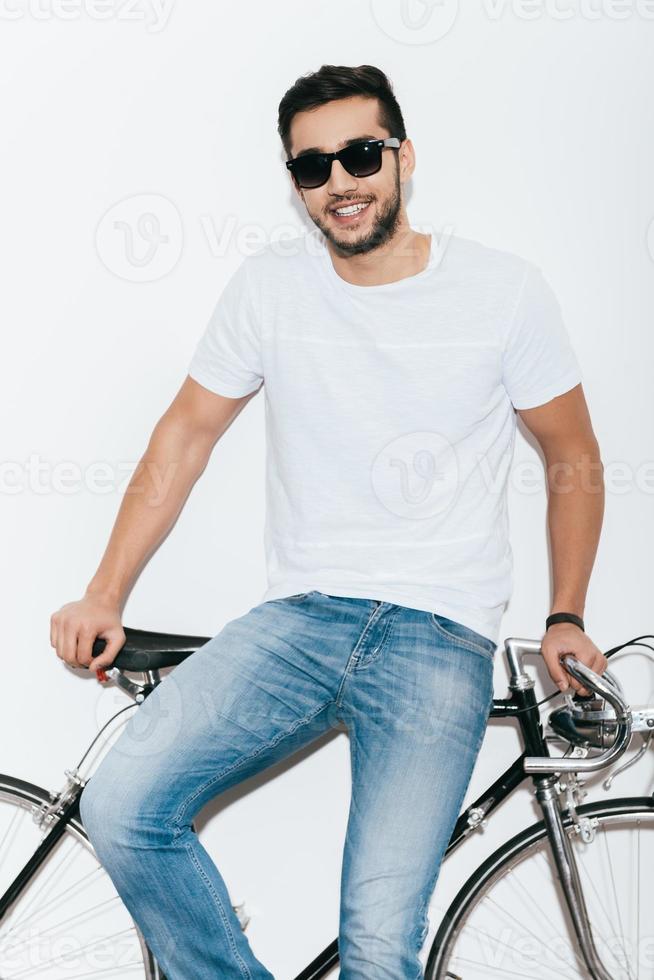 appréciant son style. beau jeune homme indien à lunettes de soleil appuyé sur son vélo de style rétro et souriant en se tenant debout sur fond blanc photo