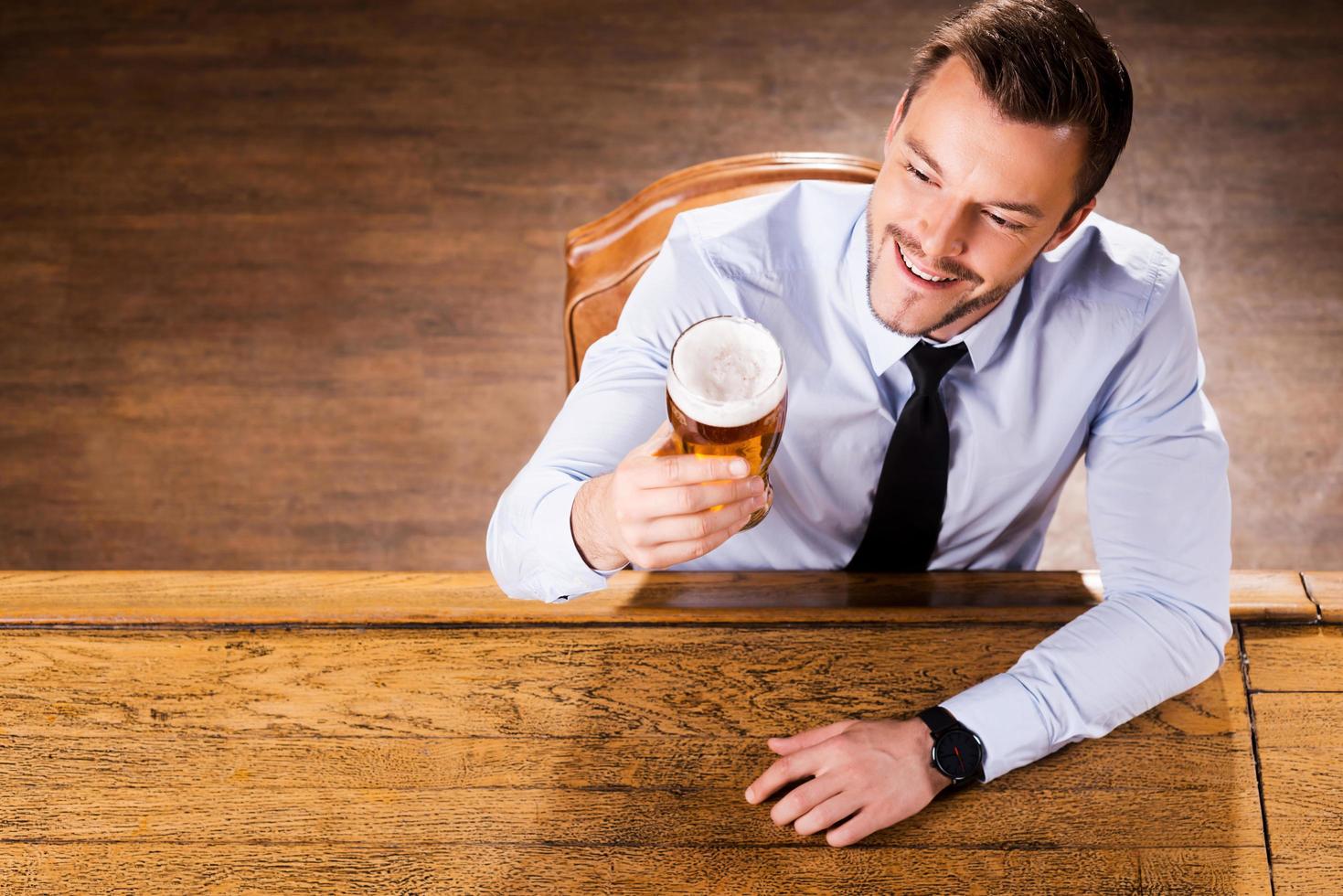 dégustant sa bière préférée. vue de dessus du beau jeune homme en chemise et cravate examinant le verre avec de la bière et souriant alors qu'il était assis au comptoir du bar photo