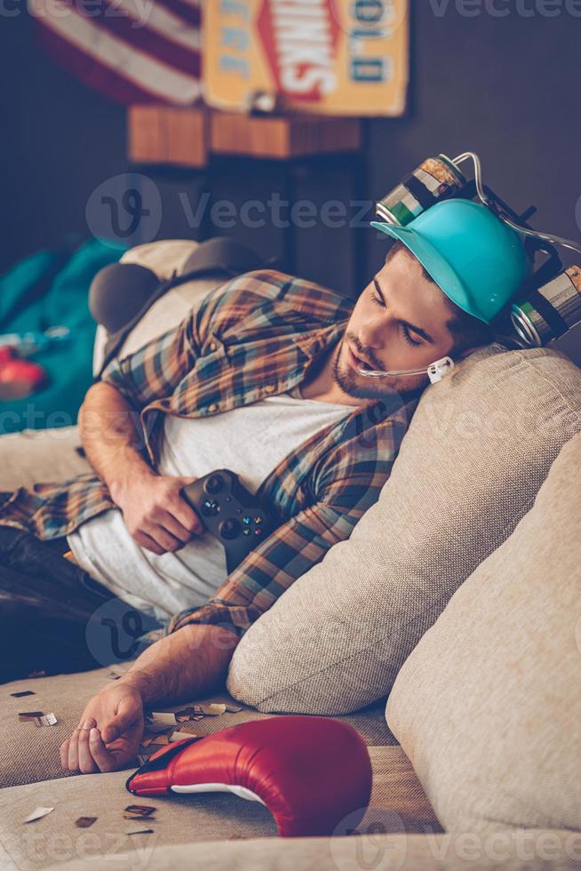 hier c'était super fête. jeune bel homme au chapeau de bière tenant le joystick et faisant la sieste sur le canapé dans une pièce en désordre après la fête photo