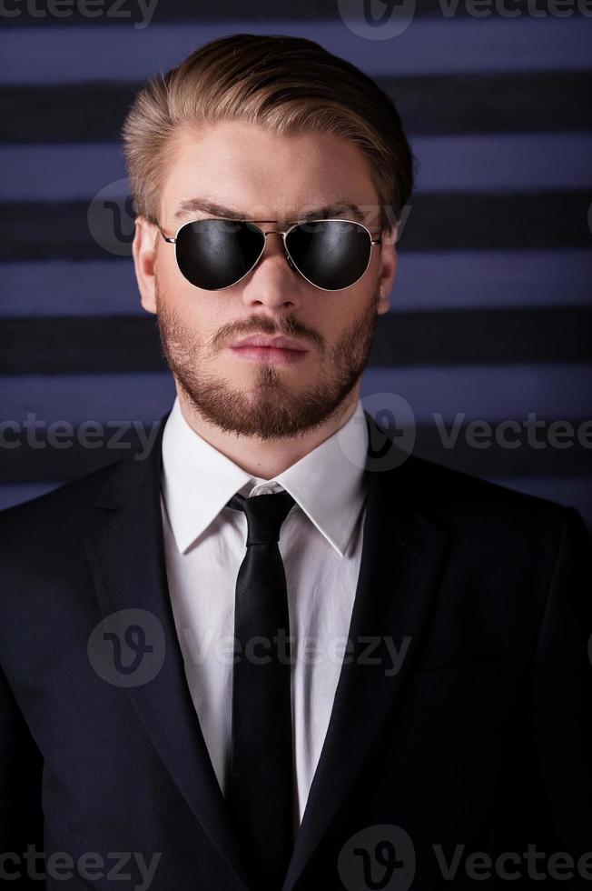 portrait de confiance et de masculinité. portrait de beau jeune homme à lunettes de soleil et tenues de soirée regardant la caméra en se tenant debout sur fond rayé photo