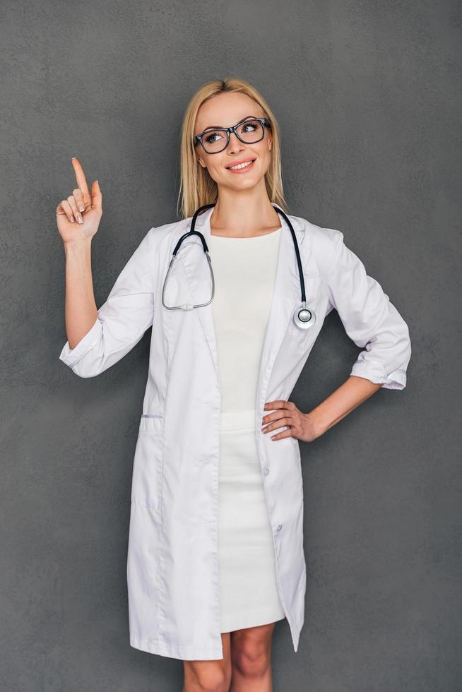 recommandation de votre médecin. belle jeune femme médecin pointant sur l'espace de copie et regardant loin avec le sourire en se tenant debout sur fond gris photo