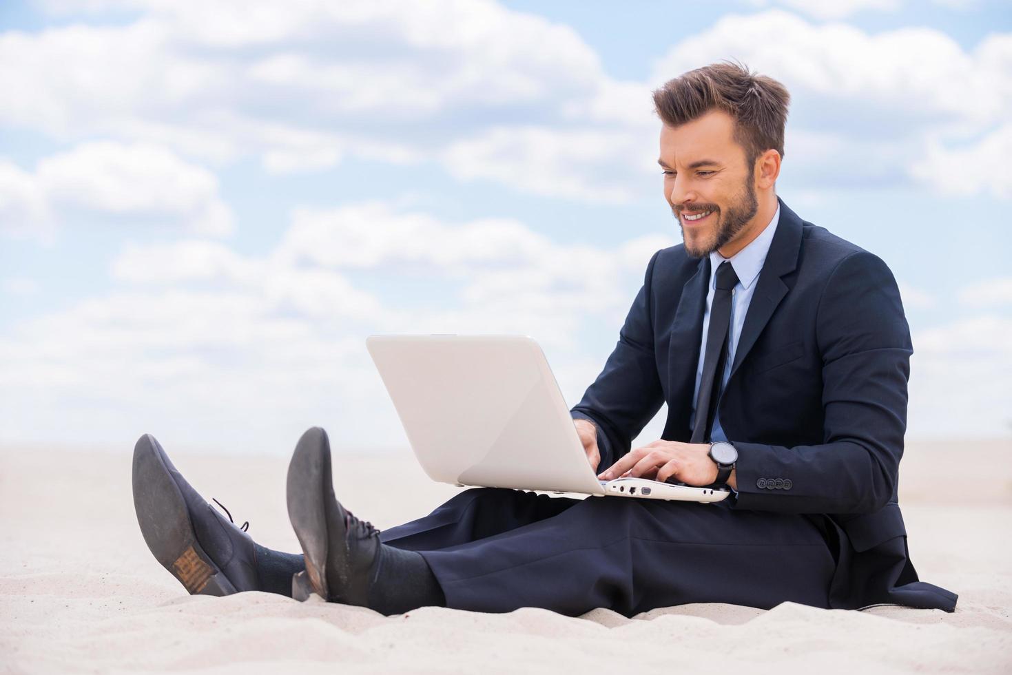 super endroit pour travailler. joyeux jeune homme en tenue de soirée travaillant sur un ordinateur portable assis sur le sable dans le désert photo