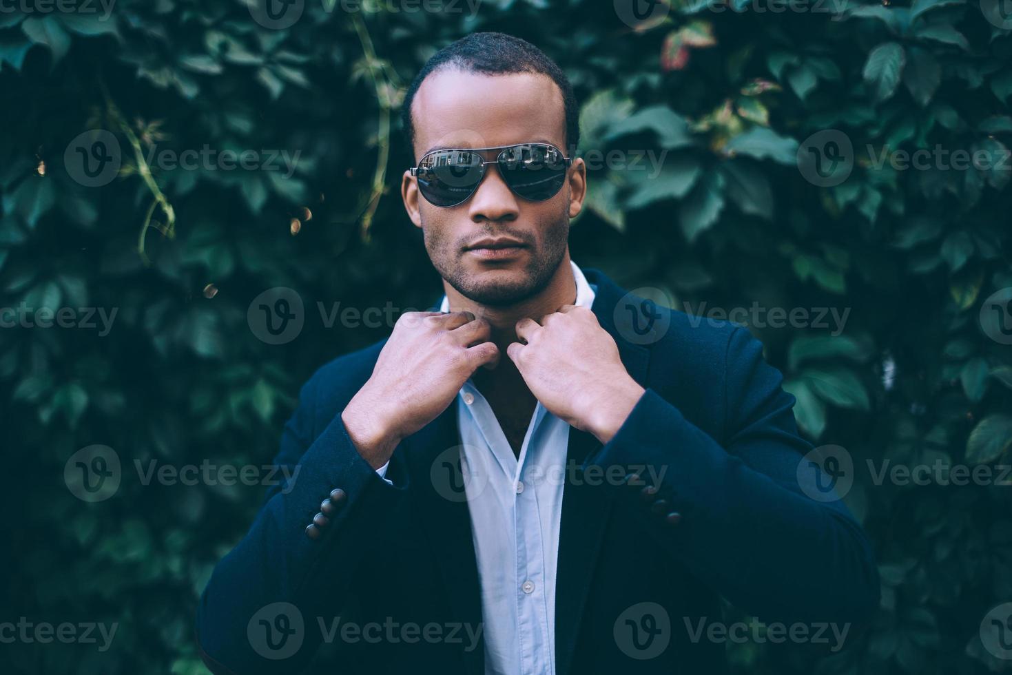 à la recherche tout simplement parfait. beau jeune homme africain en tenue décontractée intelligente ajustant son col de chemise et regardant la caméra en se tenant debout sur fond de plante verte à l'extérieur photo