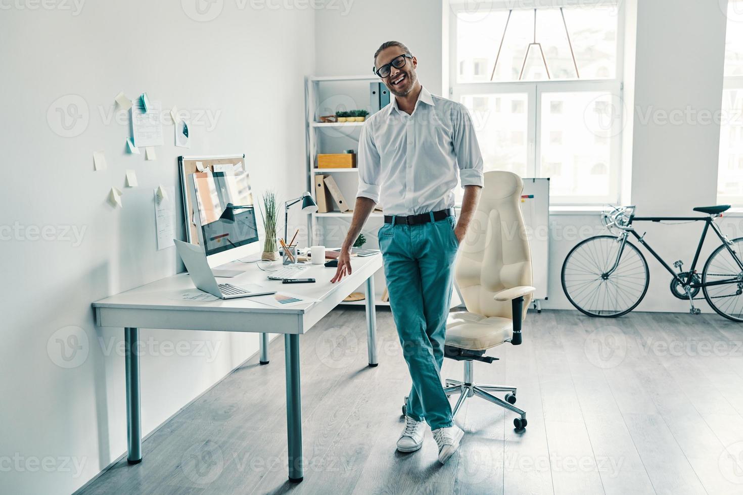 gestionnaire confiant. toute la longueur du beau jeune homme en chemise et jeans souriant tout en se tenant au bureau photo