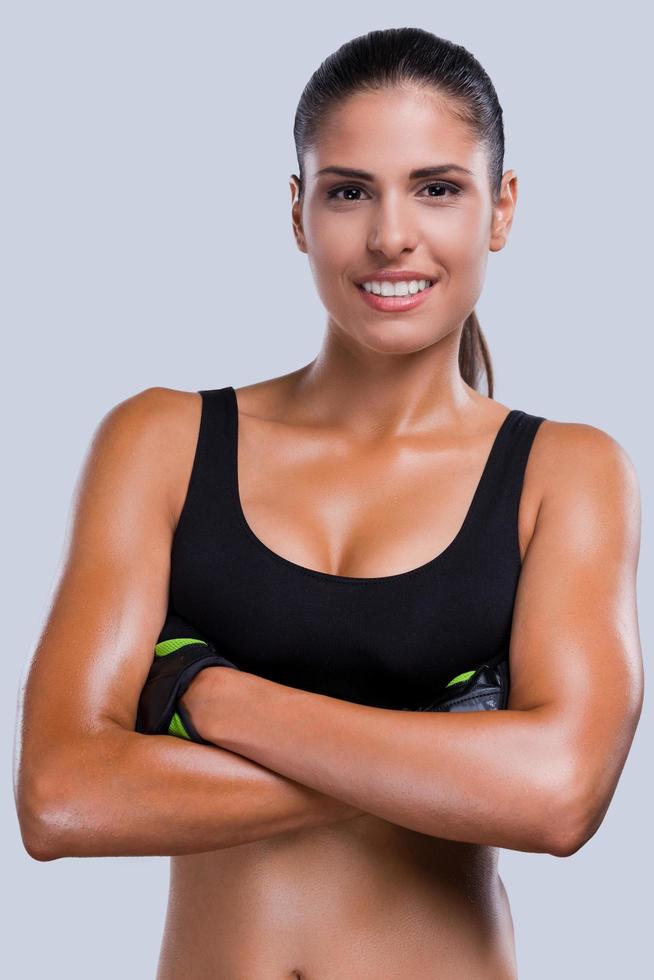 en forme et belle. belle jeune femme sportive en vêtements de sport en gardant les bras croisés et souriant en se tenant debout sur fond gris photo