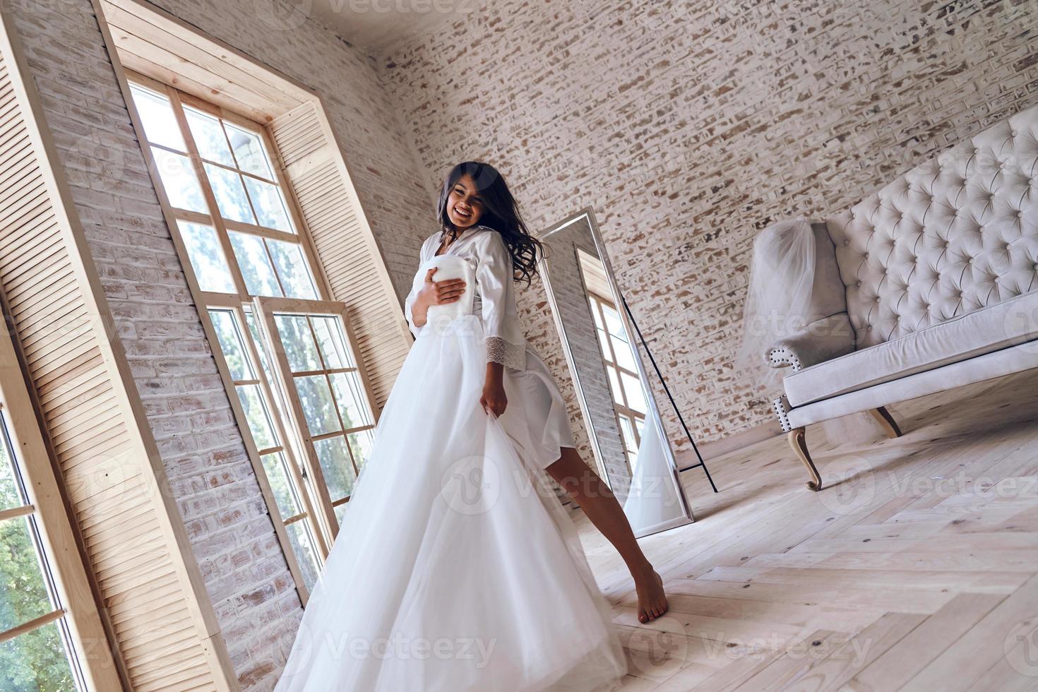 ça va être une journée magique. toute la longueur de la jolie jeune femme en peignoir de soie essayant sa robe de mariée et souriant en se tenant debout photo