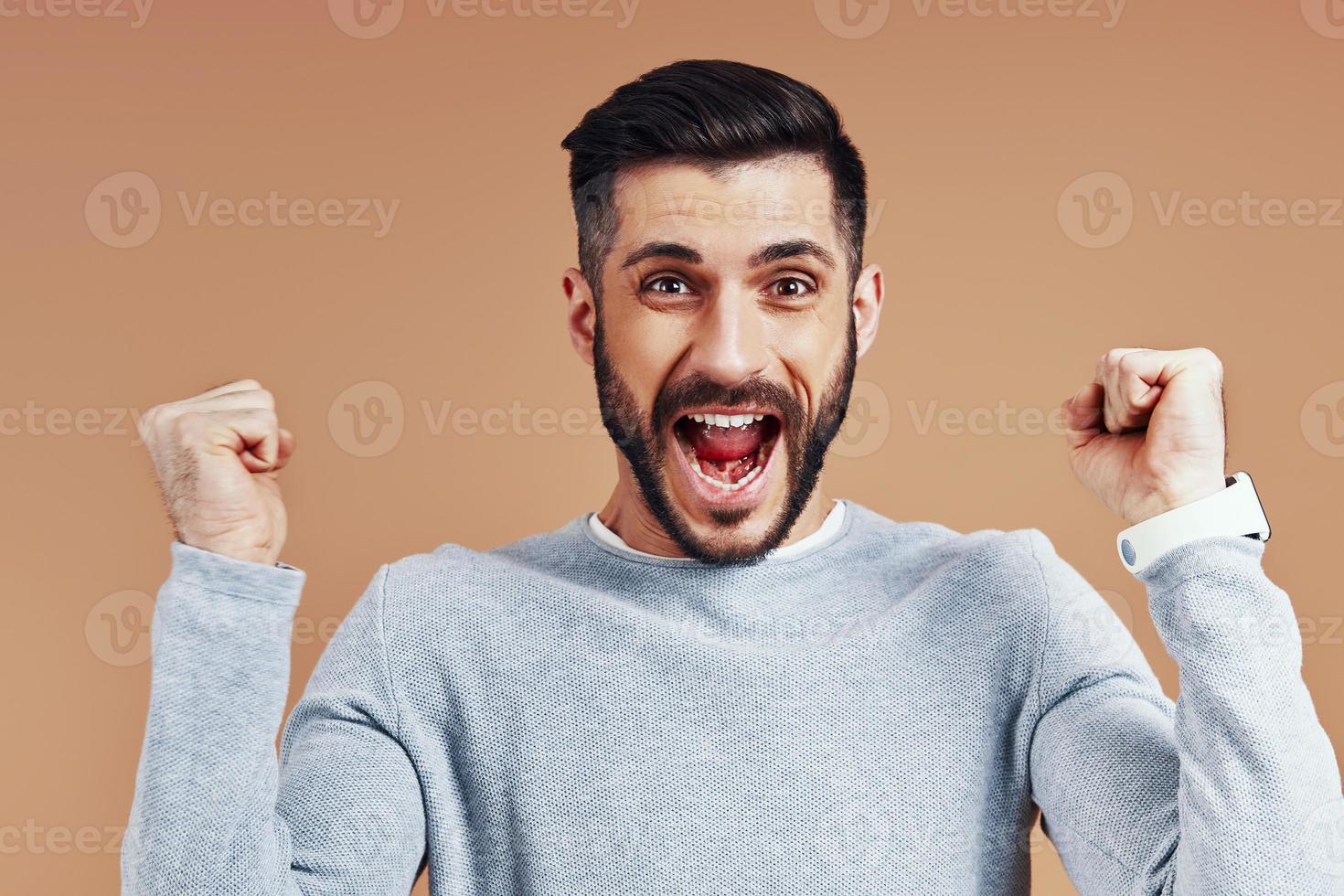 heureux jeune homme en tenue décontractée acclamant et souriant debout sur fond marron photo