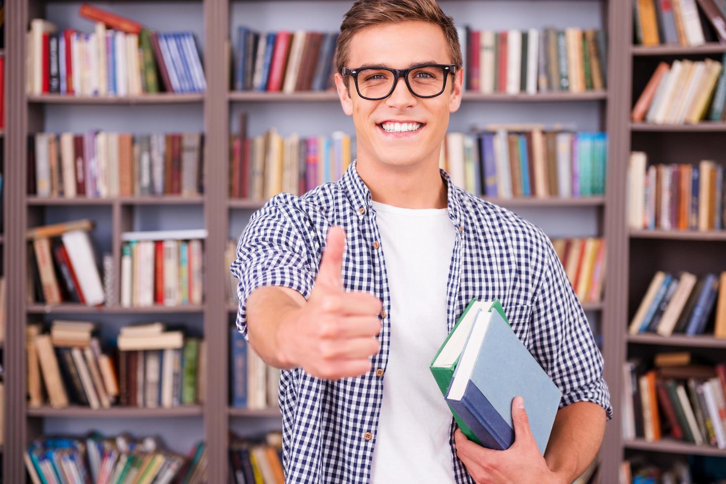 il aime étudier. beau jeune homme tenant des livres et montrant son pouce vers le haut tout en se tenant dans la bibliothèque photo