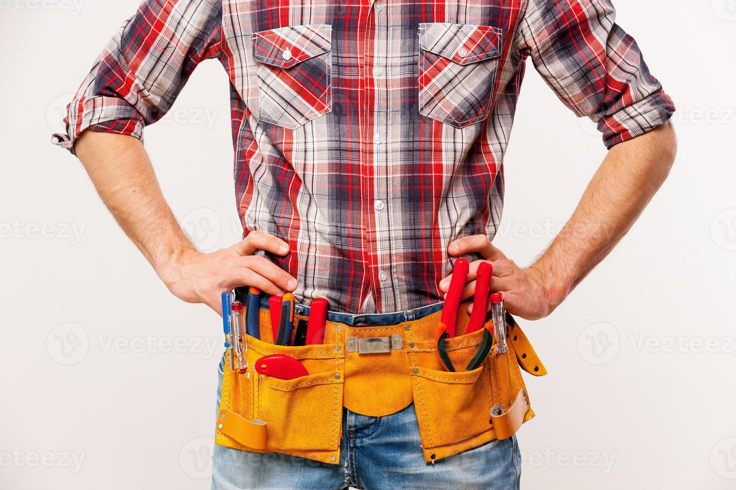 prêt à travailler. beau jeune bricoleur avec ceinture à outils tenant les mains sur la hanche en se tenant debout sur fond gris photo